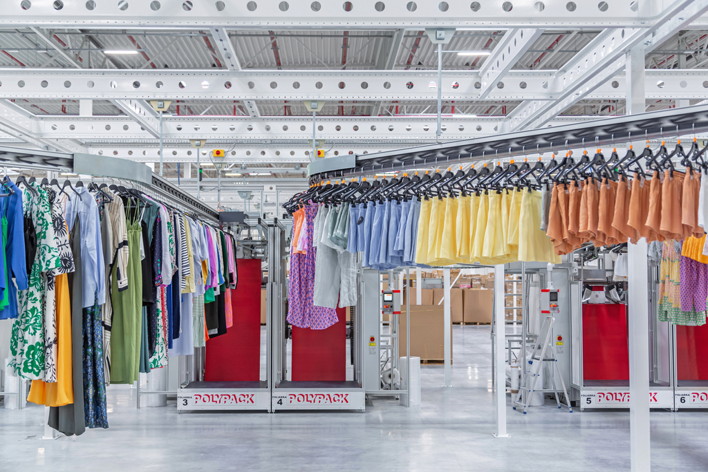 Värikäs jono vaatteita henkareissa vaatekuljettimella kulkemassa kohti pussituskonetta.