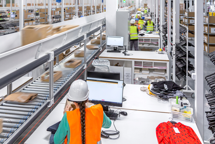 Pitkä rivi verkkokaupan palautuspöytiä valoisassa varastotilassa. Pöydillä on tuotteita, laatikoita ja tunnistuslaitteita, ja taustalla näkyy järjestetty hyllystö – kuva esittää tehokasta ja organisoitua palautusten käsittelyaluetta.